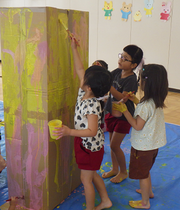 4歳児が大きな段ボールに背伸びをしながら絵の具を塗っているところ