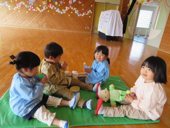 3歳児4人がマットの上に向かい合って座り、人形の赤ちゃんや飲み物を置いて飲んだり抱っこをしている姿