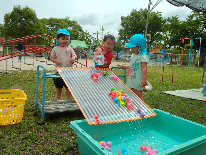 トタンに置いた魚のおもちゃを水で流している3歳児。その様子を友達が見ている。