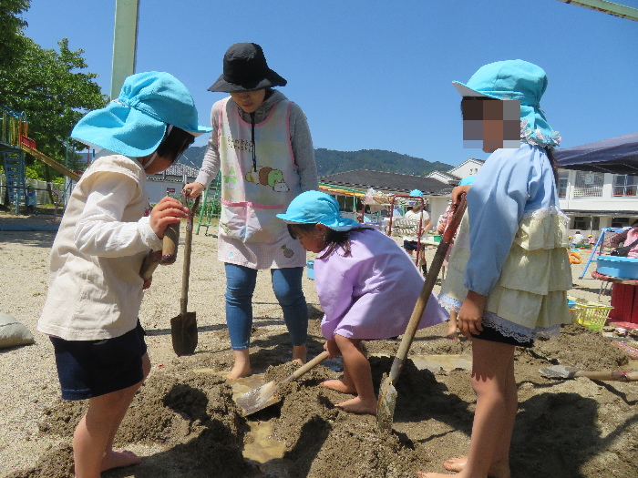 5歳児の女の子3人が先生と一緒に鉄スコップを持って砂場の砂を掘っている姿