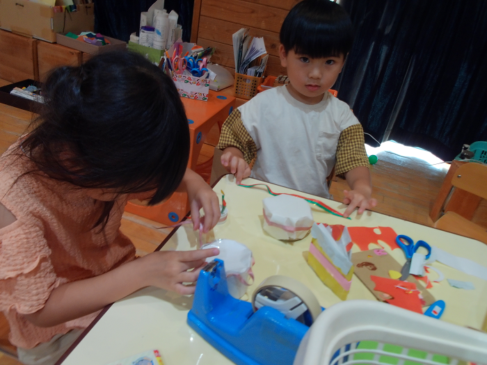 5歳児が箱やリボンを使ってケーキを作っているのを3歳児が見ている
