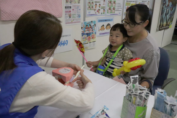 「楽しく口腔機能向上体験・ブラッシング体験コーナー」母親の膝で歯磨きのブラッシング指導を受ける男児の様子