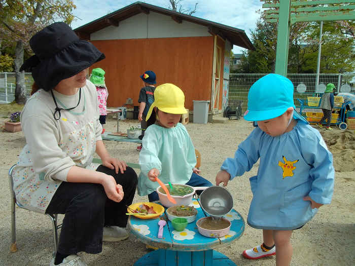 二人の園児が砂でごちそうを作って、先生に食べてもらおうとしているところ