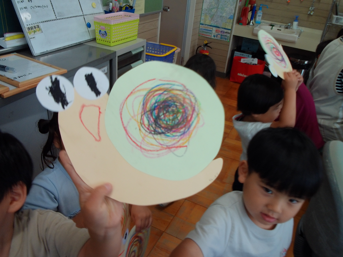 カタツムリの形の画用紙にクレパスでぐるぐる色を塗ったものを持っている3歳児