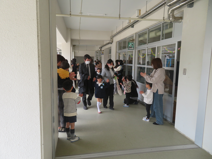 幼稚園の廊下で、卒園児が親子で歩いている両側に在園児の親子がいて、拍手で見送っている場面