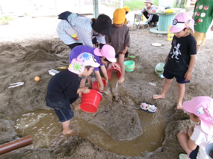 子どもたちが砂場につくった川に廃材でつくった船を水で流している様子
