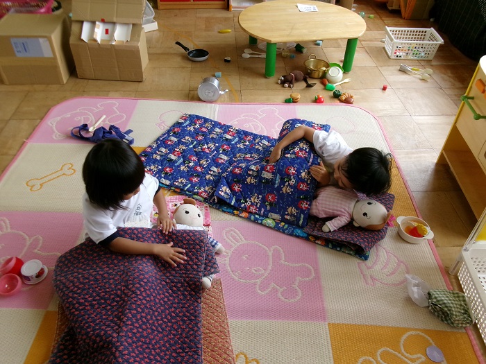 2人の子どもがおうちごっこで赤ちゃんの人形を布団に寝かせている様子
