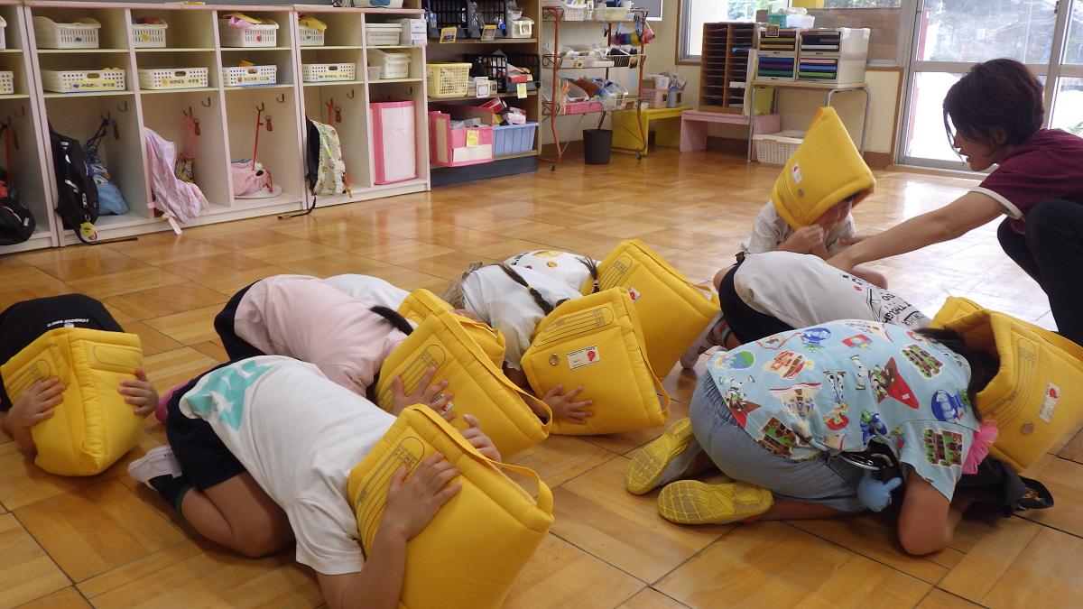 シェイクアウト訓練で防災頭巾をかぶって避難練習を行っている子どもと先生の様子